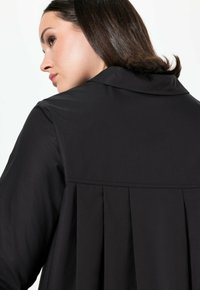 Giacca nera con un design del retro plissettato, colletto e una texture del tessuto liscia, che mette in risalto una silhouette sartoriale.