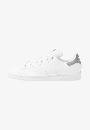Biały sneaker Adidas Stan Smith z perforowanymi paskami, białymi sznurowadłami i szarą zakładką na pięcie, przedstawiony z profilu na białym tle.