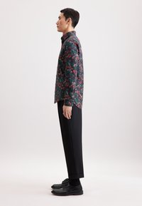 Camicia con motivo floreale in verde scuro con accenti rossi e gialli, abbinata a pantaloni neri e scarpe. Maniche lunghe, taglio dritto.