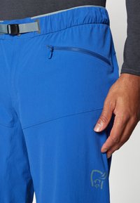 Blue hiking pants with a textured surface, zippered pocket, adjustable waistband, and a logo on the thigh. Composed of lightweight fabric.