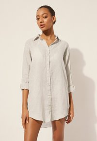 Chemise en lin gris clair avec une fermeture à boutons, manches longues retroussées et un col. Texture lisse et ourlet arrière légèrement plus long.
