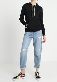 Persona vistiendo una sudadera con capucha negra, jeans rotos de color azul claro con dobladillos enrollados y zapatillas negras con diseños de llamas rojas y amarillas.