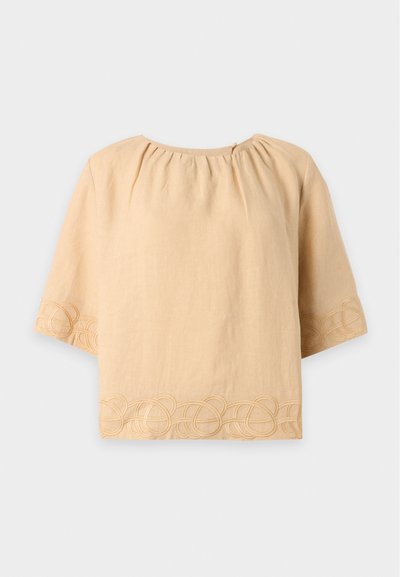 Blouse beige à manches courtes avec encolure ronde et motif de boucles brodées le long de l’ourlet et des bords des manches.