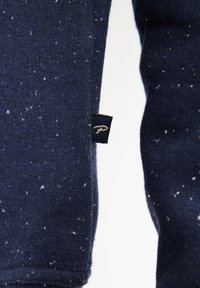 Marineblauer Strickstoff mit weißen Sprenkeln, versehen mit einem kleinen schwarzen Etikett mit einem auf die Seite gestickten Logo. Glatte Textur und figurnahe Form.