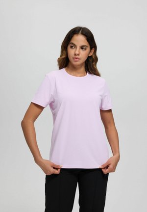 Woman with wavy hair wearing a light purple short-sleeve t-shirt and black pants, holding the shirt hem, looking to her right.