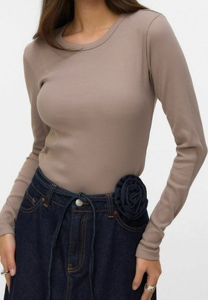 Kobieta ubrana w dopasowany, beżowy, prążkowany top z długim rękawem, włożony do wysokich, ciemnych dżinsów z materiałowym kwiatem przymocowanym do talii.