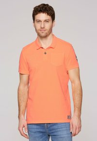 Camisa polo coral con cuello, que presenta un bolsillo en el pecho, detalles bordados y mangas cortas. Llevada con jeans azules.
