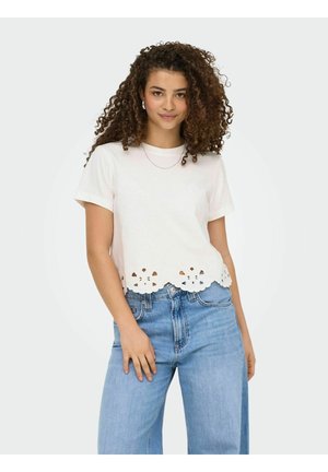 Młoda kobieta o kręconych włosach, ubrana w biały krótki top z kwiatowymi wycięciami na dole oraz jasnoniebieskie, wysokie jeansy.