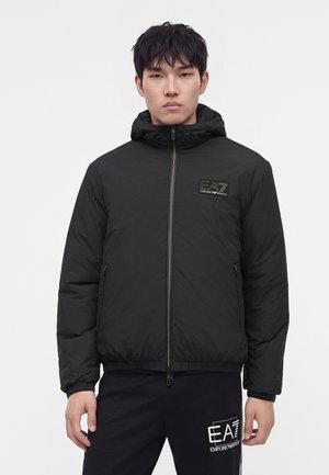 Joven con una chaqueta acolchada negra Emporio Armani EA7 con capucha y pantalones negros a juego, de pie frente a un fondo blanco liso.