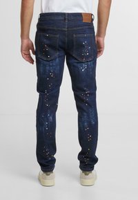 Tmavě modré džíny s úzkým střihem, dekorované barevnými skvrnami barvy a dvěma zadními kapsami. Hnědá kožená nášivka na pásu.