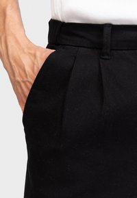 Black trousers made of textured fabric, featuring a pleated design, a classic waistband, and a side pocket with a hand inserted.