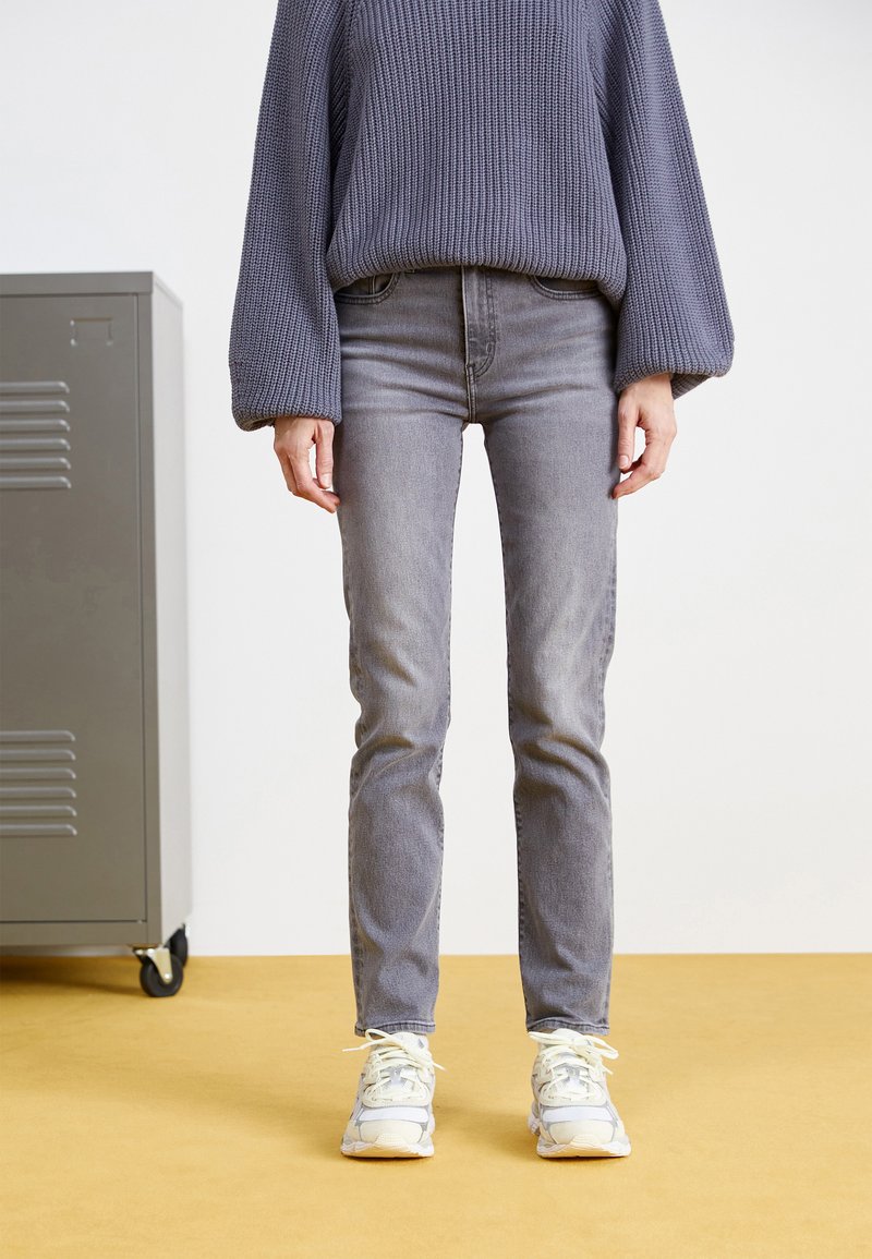 Jeans grigi con una vestibilità slim, realizzati in denim, abbinati a un maglione grigio morbido e testurizzato e sneakers bianche. Lo sfondo è un pavimento giallo uniforme.