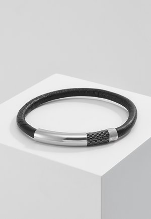 Bracelet en cuir noir avec une fermeture en métal argenté et noir texturé, présenté sur un socle blanc sur un fond gris.