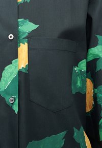 Σκούρο πράσινο πουκάμισο με μεγάλο κίτρινο και πράσινο φύλλωμα, τσέπη στο μπροστινό μέρος στο στήθος και ορατά κουμπιά κατά μήκος της πατιλέτας.