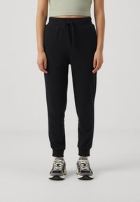 Schwarze Jogginghose mit elastischem Bund und Kordelzug, ausgestattet mit einem schmal zulaufenden Bein-Design. Kombiniert mit gemusterten Sportschuhen.