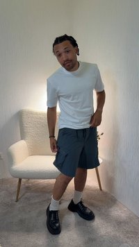 Homme aux cheveux tressés portant un t-shirt blanc, un short cargo sombre, des chaussettes blanches et des chaussures noires, se tenant près d'un fauteuil blanc texturé dans une pièce avec moquette.
