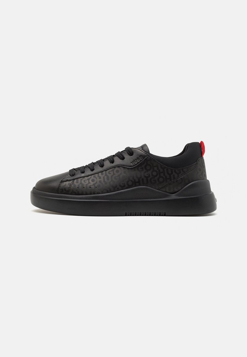 Baskets basses noires avec motif en relief du nom de la marque répété, lacets noirs, semelle épaisse et languette rouge au talon sur fond blanc.