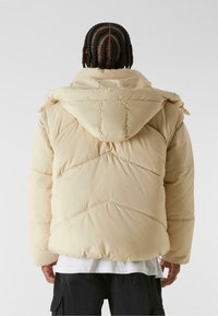 Puffer jacket in light beige with a hood, featuring a quilted design and contrasting stitching. Soft, padded texture.
