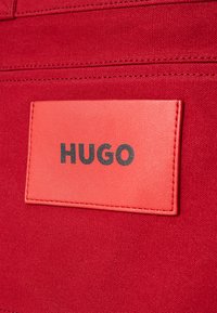 Červená textilná tkanina s obdĺžnikovou červenou koženou nášivkou, na ktorej je v čiernej farbe embosovaná značka "HUGO". Okolo nášivky sú viditeľné prešívané detaily.