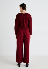 Red, textured sweater with a wide neckline, paired with high-waisted, wide-leg trousers in matching red fabric. Black heeled boots.