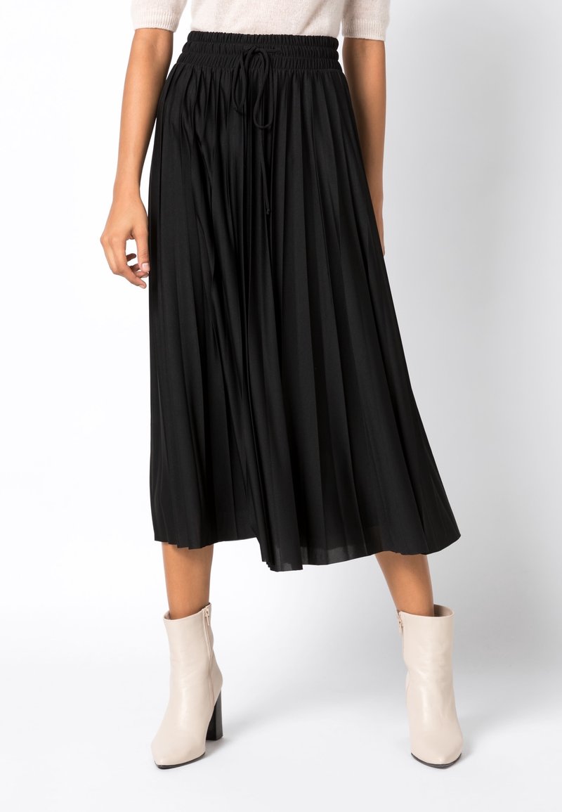 Femme portant une jupe mi-longue plissée noire avec une taille élastique et des bottines crème, sur un fond uni.