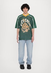 Zöld hálós, oversized póló, sárga nyakkal és fehér csíkokkal, egy fagylaltot tartó karakter grafikájával díszítve.