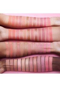 Blushs in verschillende tinten roze en perzik op armen, met matte en glanzende afwerkingen, tegen een roze achtergrond.