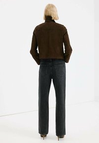 Veste courte en daim marron avec un col montant, associée à un jean gris foncé taille haute et des talons aiguilles beiges, vue de dos.