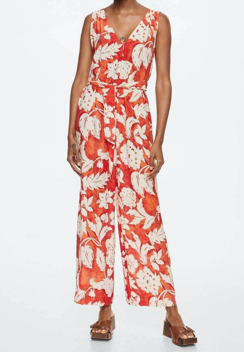 Femme portant une combinaison orange sans manches avec un grand motif floral blanc et des sandales compensées marron sur fond blanc uni.
