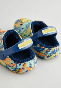 Des sabots colorés sur le thème de Pokémon avec un intérieur en peluche. Ils disposent d'une sangle bleue étiquetée "POKÉMON" et d'un extérieur texturé et à motifs.
