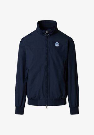 Veste zippée bleu marine avec col montant, poignets et ourlet élastiques, poches latérales zippées, et logo circulaire sur la poitrine gauche.
