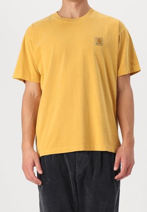 Mand iført en enkel gul kortærmet T-shirt med lille Carhartt-logo på brystet og mørke fløjlsbukser, stående mod en hvid baggrund.