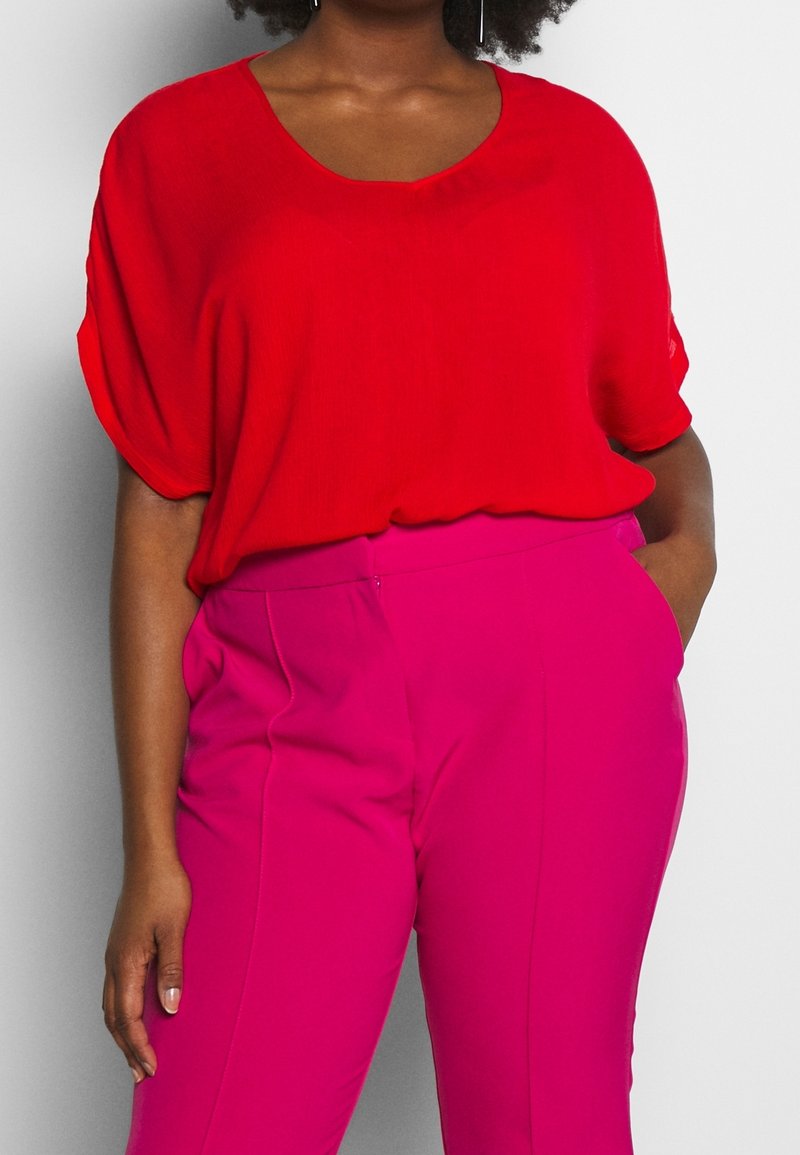 Haut rouge surdimensionné avec un col en V et des manches courtes, associé à un pantalon rose taille haute, présentant une texture lisse et une coupe ajustée.
