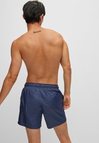 Sötétkék fürdőshorts elasztikus derékkal, oldalsó zsebekkel és egy kis piros díszítéssel. Simább, könnyű anyag, egyszerű dizájnnal.