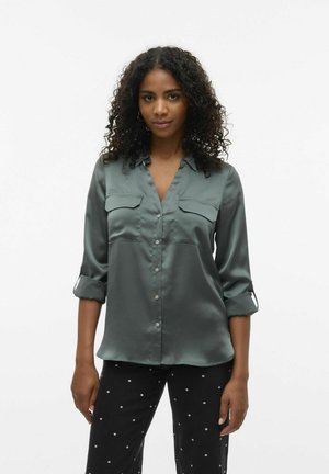 Blouse en satin vert avec un col, deux poches poitrine et des manches retroussées. Le tissu a une texture lisse avec une finition brillante.