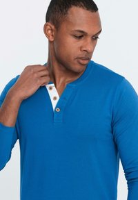 Maglietta a maniche lunghe henley blu in cotone, con una patta di bottoni con accenti crema e bottoni di legno. Texture liscia.