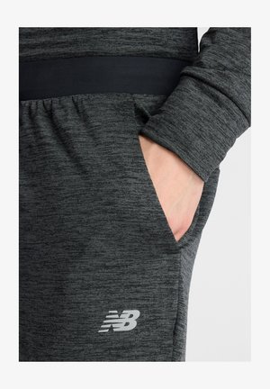 Dark grey athletic pants with a soft texture, elastic waistband, and side pockets. Features a reflective "NB" logo on the lower leg.