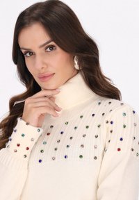 Strikpullover i creme med en ribbet turtleneck. Har flerfarvede juveldetaljer langs skuldrene og manchetterne.