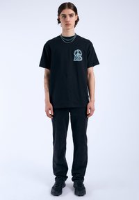 Camiseta de algodón negra con gráfico azul en el lado izquierdo del pecho, combinada con pantalones negros y zapatillas negras. Diseño sencillo con corte relajado.
