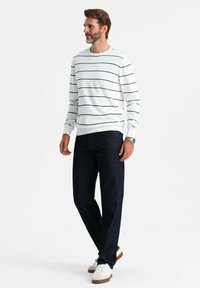 Camisa blanca de manga larga con rayas horizontales en azul marino, combinada con jeans de mezclilla oscuros y zapatillas blancas, con un corte relajado y cuello redondo.