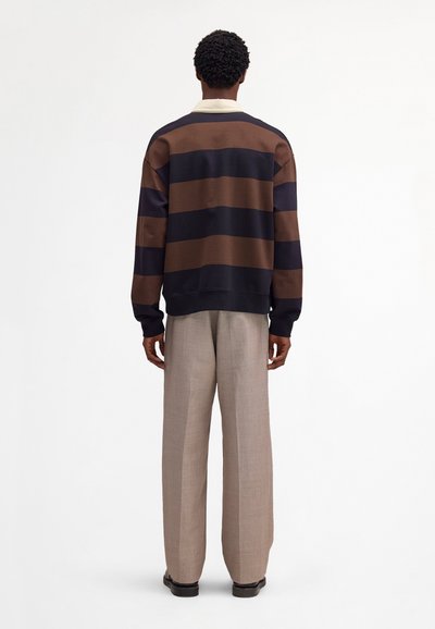 Homme debout, de dos, portant une chemise à manches longues à larges rayures marron et noires, un pantalon à jambes larges beige et des chaussures noires.