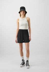 Tricou alb fără mâneci, pantaloni scurți negri cu șnur, pălărie neagră tip bucket cu dungi albe, șosete albe și teniși negri cu glezna înaltă.