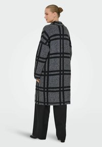Długi, oversized płaszcz w kolorze szarym z czarnym wzorem w kratę, wykonany z teksturowanej mieszanki wełny. Charakteryzuje się wysokim kołnierzem i długimi rękawami.