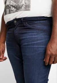 Dark blue denim jeans with a slim fit, featuring a front pocket, metal button closure, and subtle texture lines.