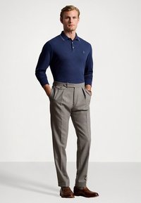 Tengerészkék hosszú ujjú galléros póló két gombbal, világosszürke pliszírozott nadrággal oldalrögzítő fülekkel, és barna bőr cipővel párosítva.