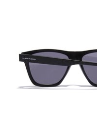 Ochelari de soare cu ramă din acetat negru, formă unghiulară, lentile închise la culoare și logo-ul argintiu "HAWKERS" pe bradă. Textură netedă și design modern.