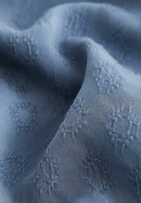 Tissu bleu à texture douce, présentant un motif géométrique répétitif subtil. Le matériau semble léger et lisse.
