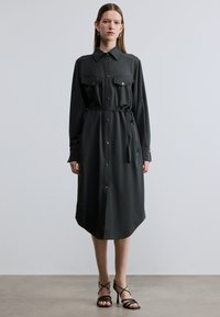 Σκούρο πράσινο φόρεμα με κουμπιά, μακριά μανίκια, δύο τσέπες στο στήθος, ζώνη στη μέση και καμπυλωτό τελείωμα. Λείο ύφασμα με ελάχιστη γυαλάδα.