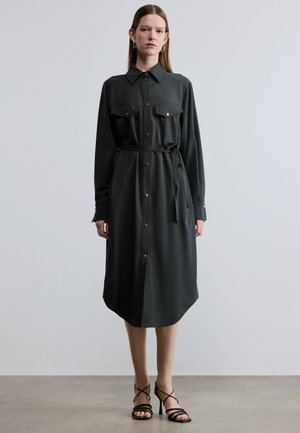 Donkergroene blousejurk met lange mouwen, twee borstzakken, een taille met riem en een afgeronde zoom. Glad stof met minimale glans.