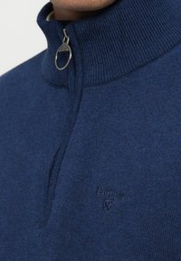 Camisola azul-marinho com fecho de correr no pescoço, feita de tecido de malha macia. Apresenta uma gola alta, acabamentos canelados e um discreto logótipo bordado no peito.